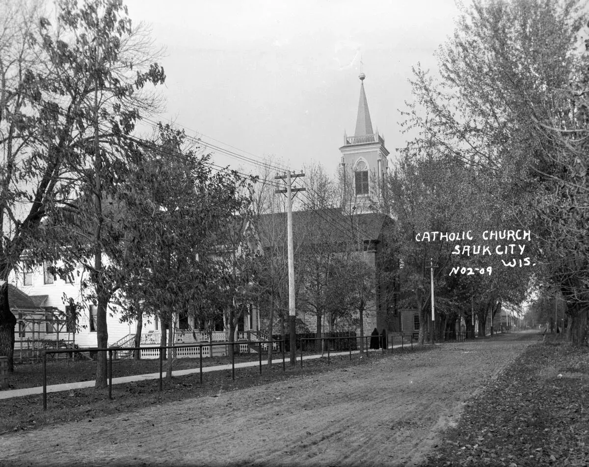 A church with a tall steeple behind trees on an unpaved street in Sauk City, 1909