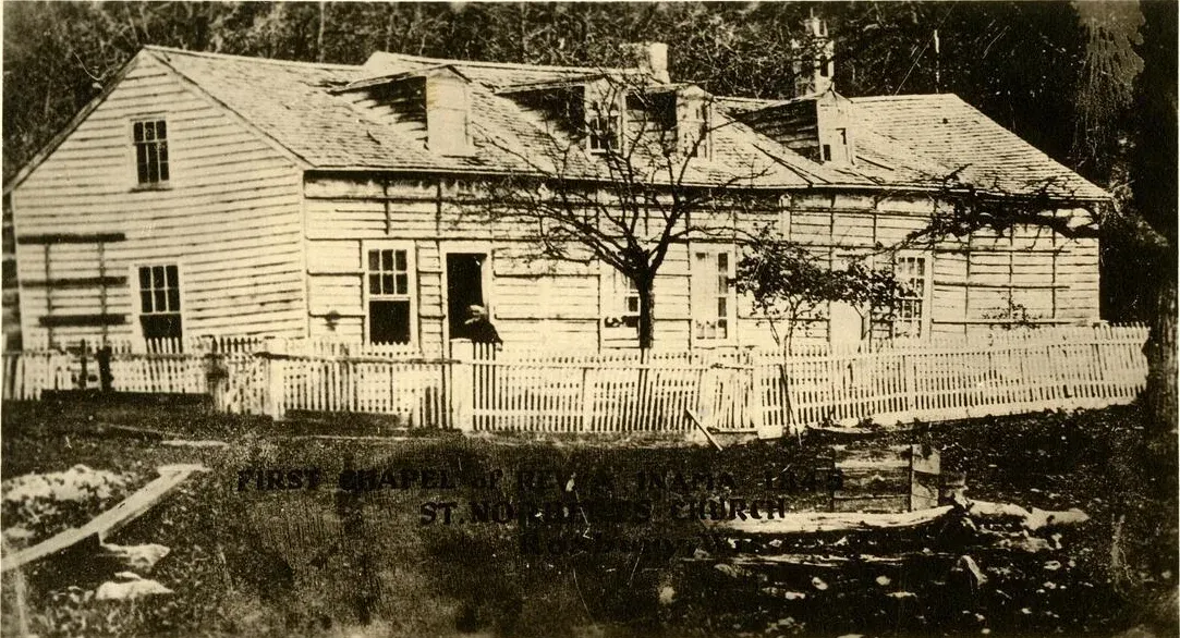 A long frame house behind a picket fence, captioned as Fr. Inama’s first chapel